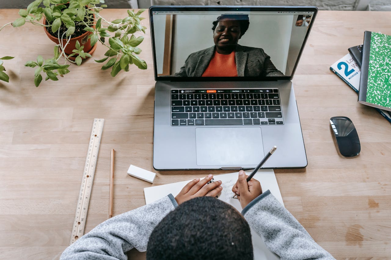 A student takes notes during an online class with a teacher on a video call.